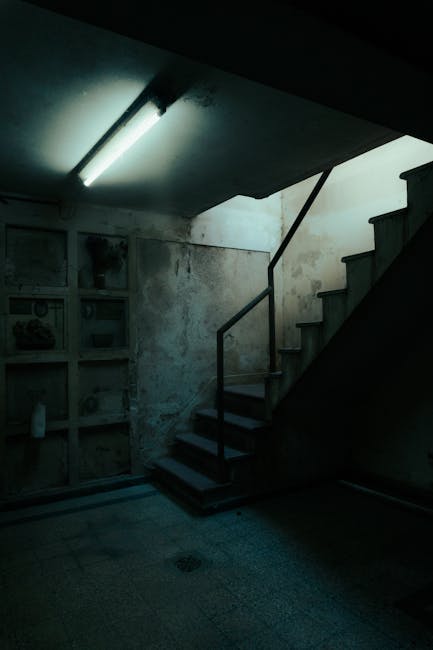 Interior view of a stairwell in a residential building, illuminated by a single fluorescent ceiling light. The staircase has dark wooden steps with a simple metal handrail on the right side, leading u