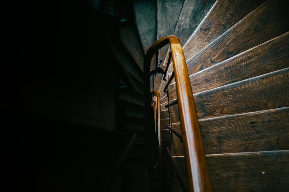 Interior view of a stairwell in a residential building, illuminated by a single fluorescent ceiling light. The staircase has dark wooden steps with a simple metal handrail on the right side, leading u